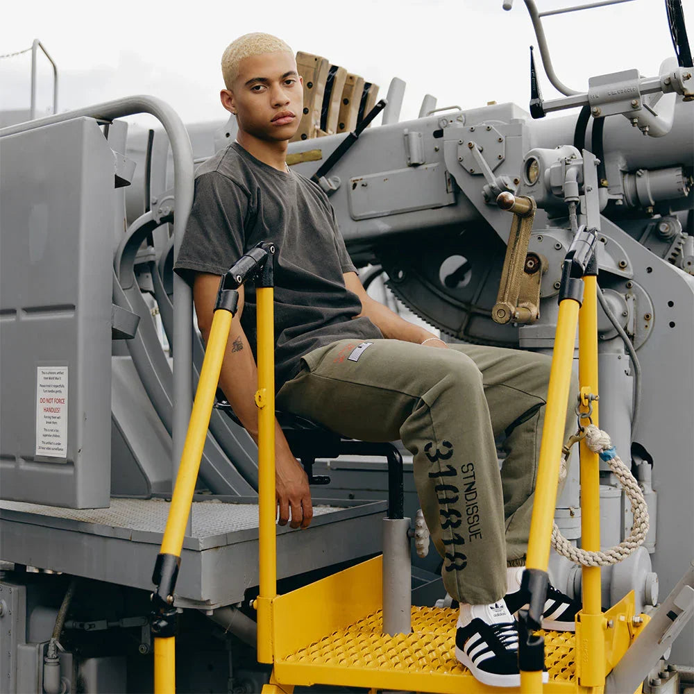 Man wearing the burnt olive Eminem Kamikaze x Standard Issue sweatpants seated on deck