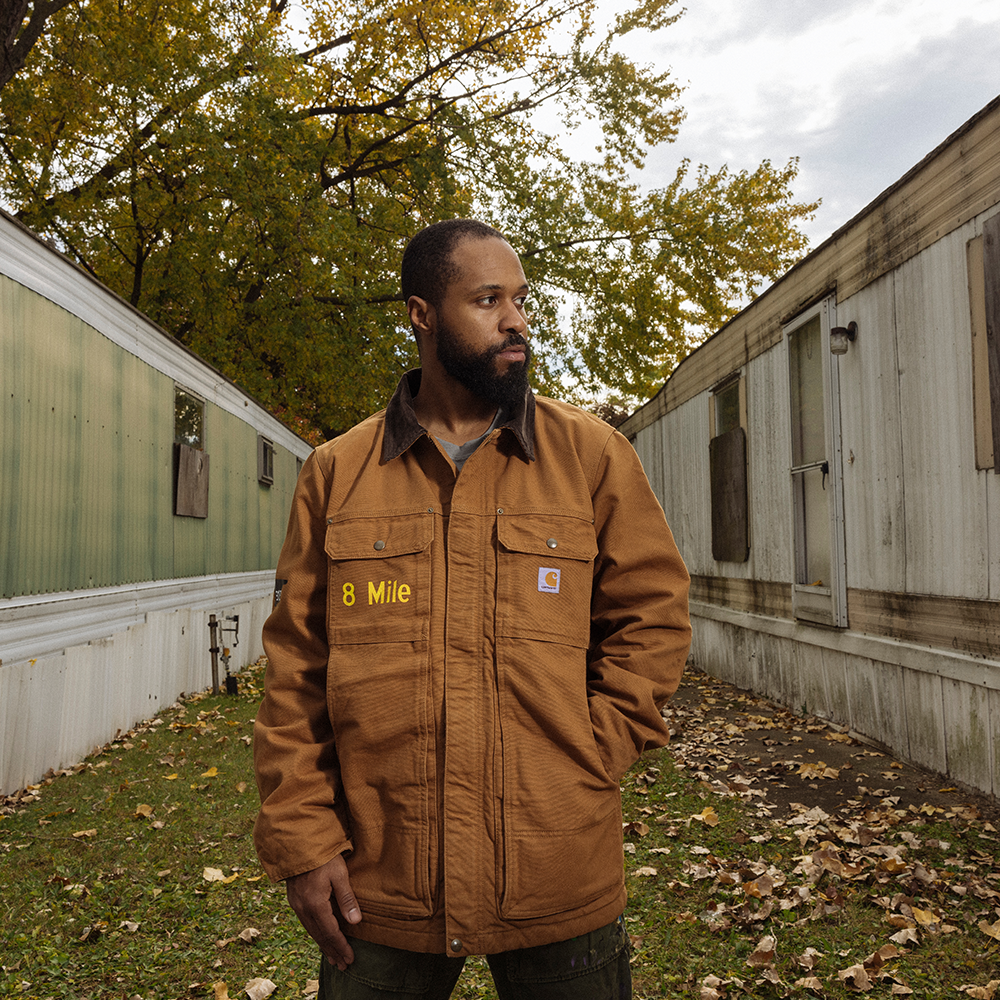 Man wearing brown Eminem 8 Mile Carhartt jacket outdoors beside a Detroit trailer home