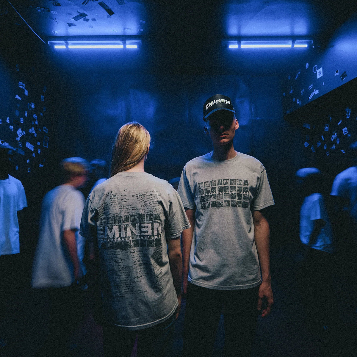 Two models in grey Eminem MMLP25 Scribble T-shirts in a blue-lit crowd