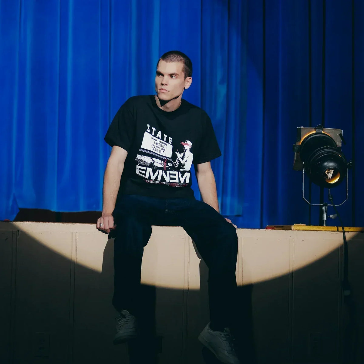 Model seated in the black Eminem Encore Dynamite T-shirt on a blue-curtained stage