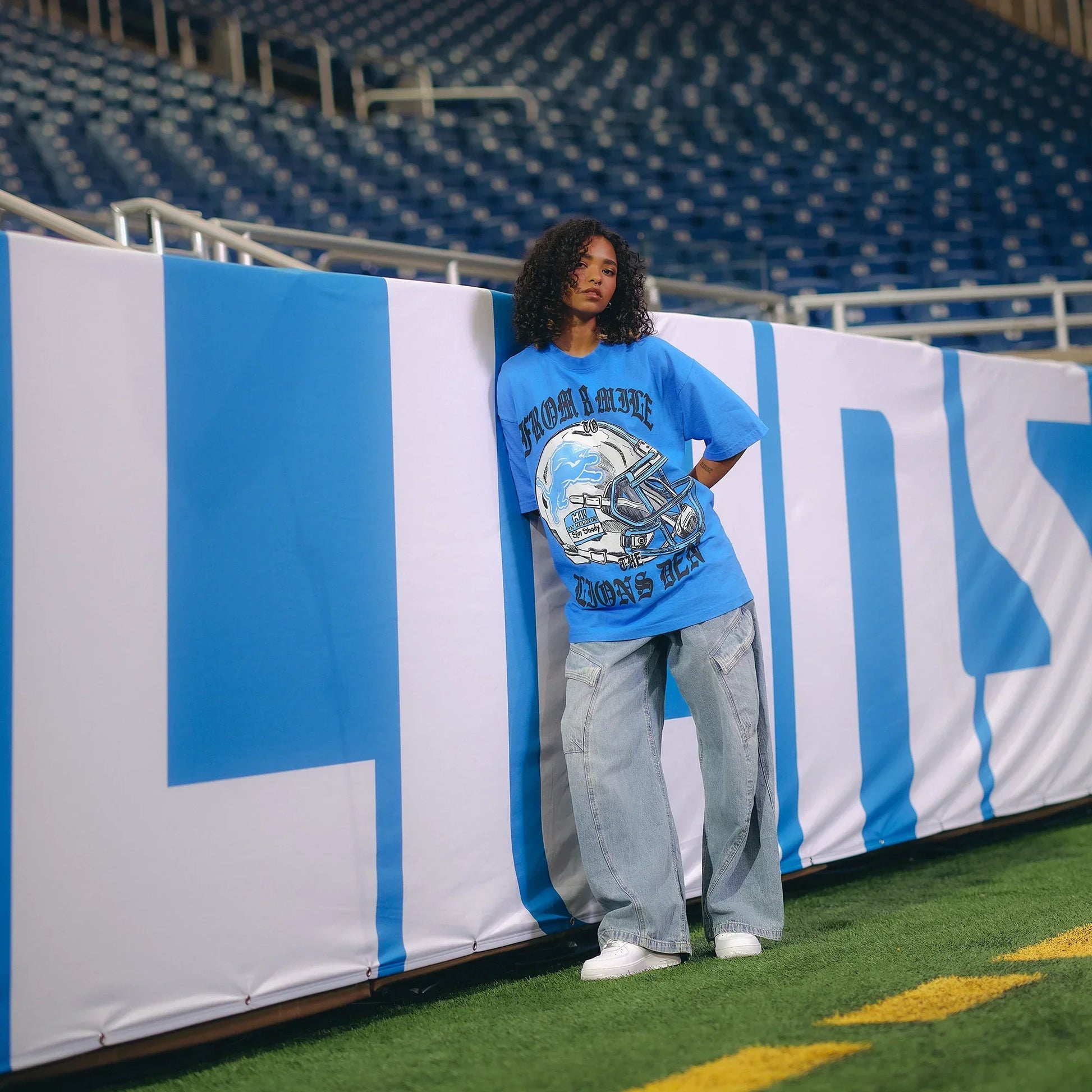 Woman in the blue Eminem x Detroit Lions Helmet T-shirt beside a Lions banner