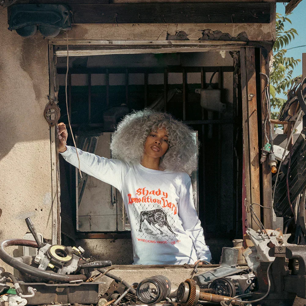 Woman wearing the white Eminem Junkyard Dog long sleeve in a scrapyard doorway